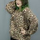 A woman modeling a leopard print long-sleeve sweater and black pants with side grommets, standing against a gray studio backdrop.