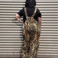 A person standing with their back to the camera in front of a metal roll-up garage door, wearing leopard-print overalls over a black t-shirt and black shoes.