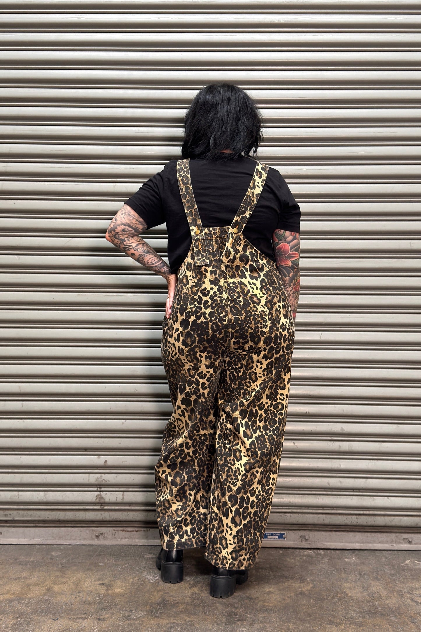 A person standing with their back to the camera in front of a metal roll-up garage door, wearing leopard-print overalls over a black t-shirt and black shoes.