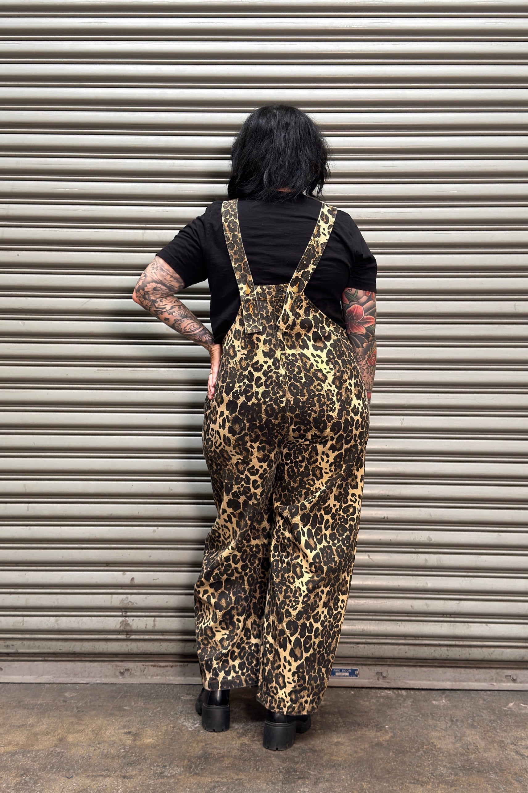A person standing with their back to the camera in front of a metal roll-up garage door, wearing leopard-print overalls over a black t-shirt and black shoes.
