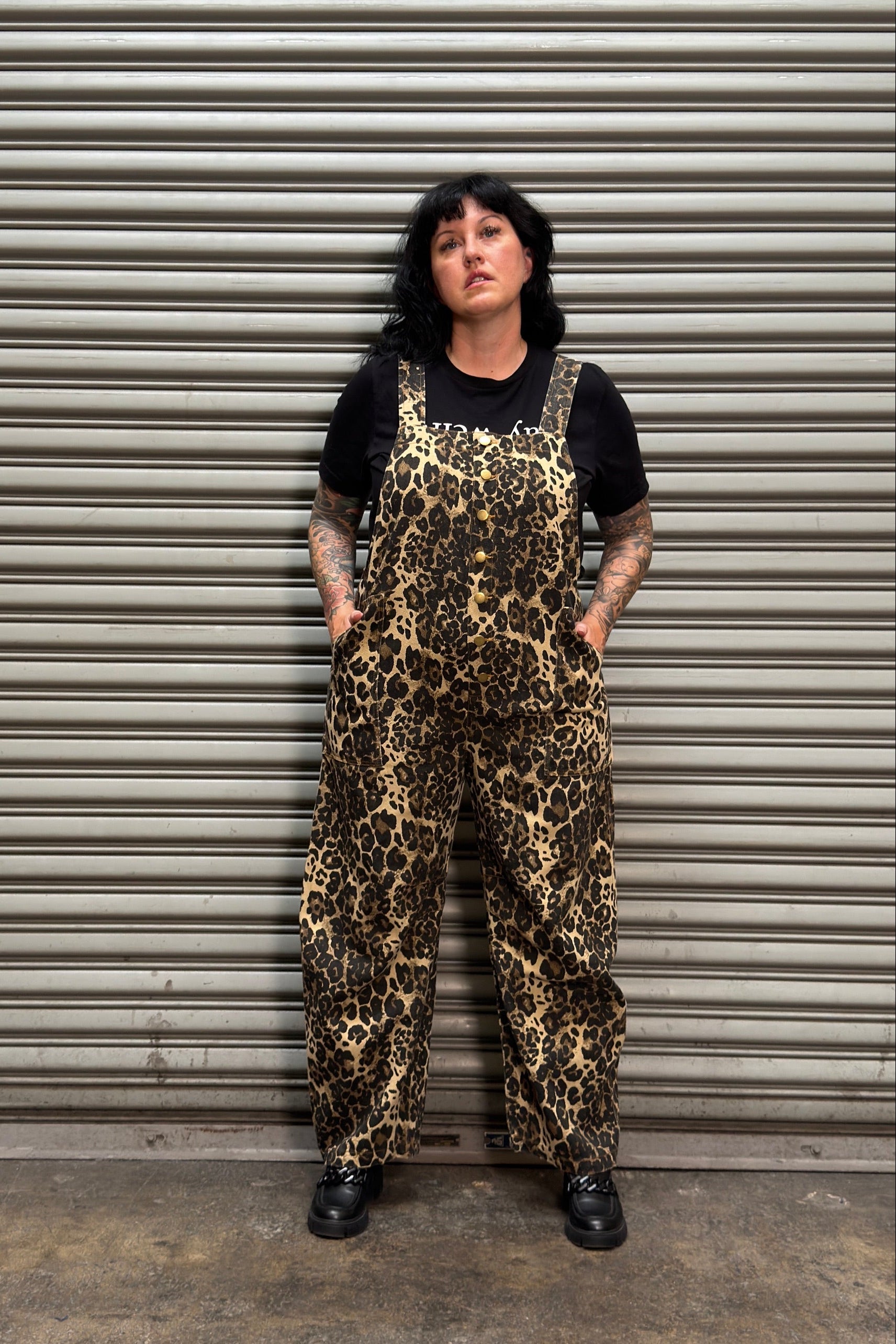 Women with black hair and tattoos standing in front of metal backdrop wearing leopard overalls.