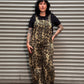 Woman with black hair and tattoos standing in front of a metal backdrop wearing leopard print overalls.