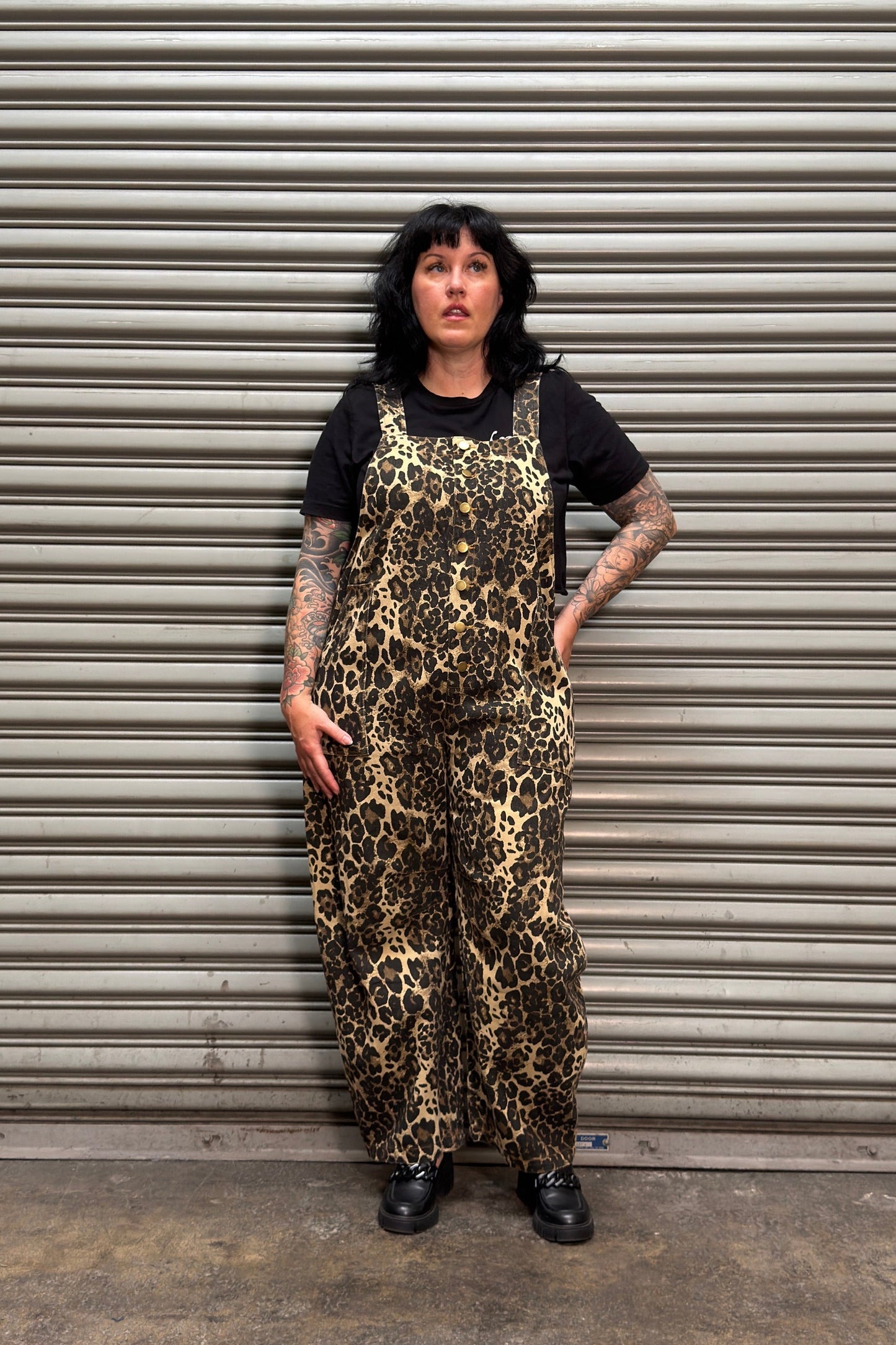 Woman with black hair and tattoos standing in front of a metal backdrop wearing leopard print overalls.