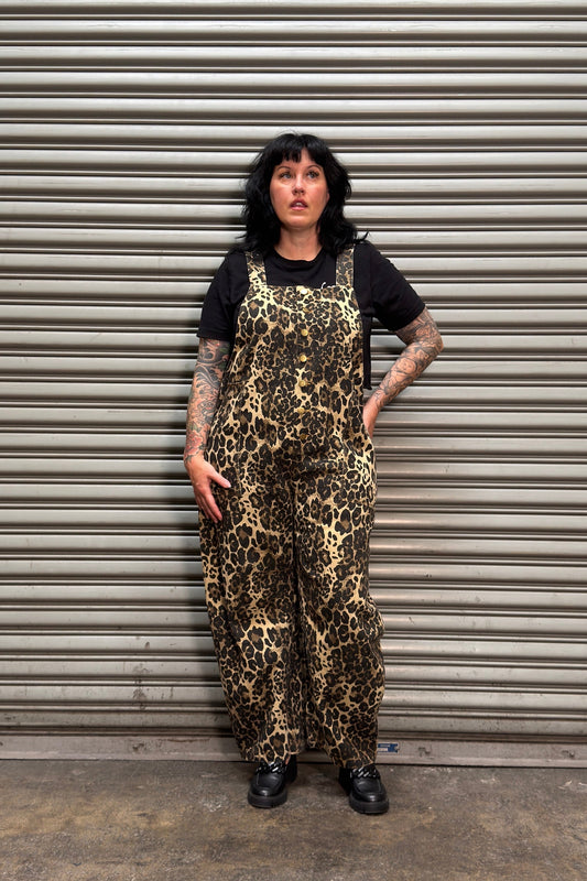 Woman with black hair and tattoos standing in front of a metal backdrop wearing leopard print overalls.