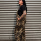 A person with dark hair and visible arm tattoos stands in profile in front of a gray metal roll-up door, wearing leopard-print overall-style shorts over a black T-shirt and black shoes.