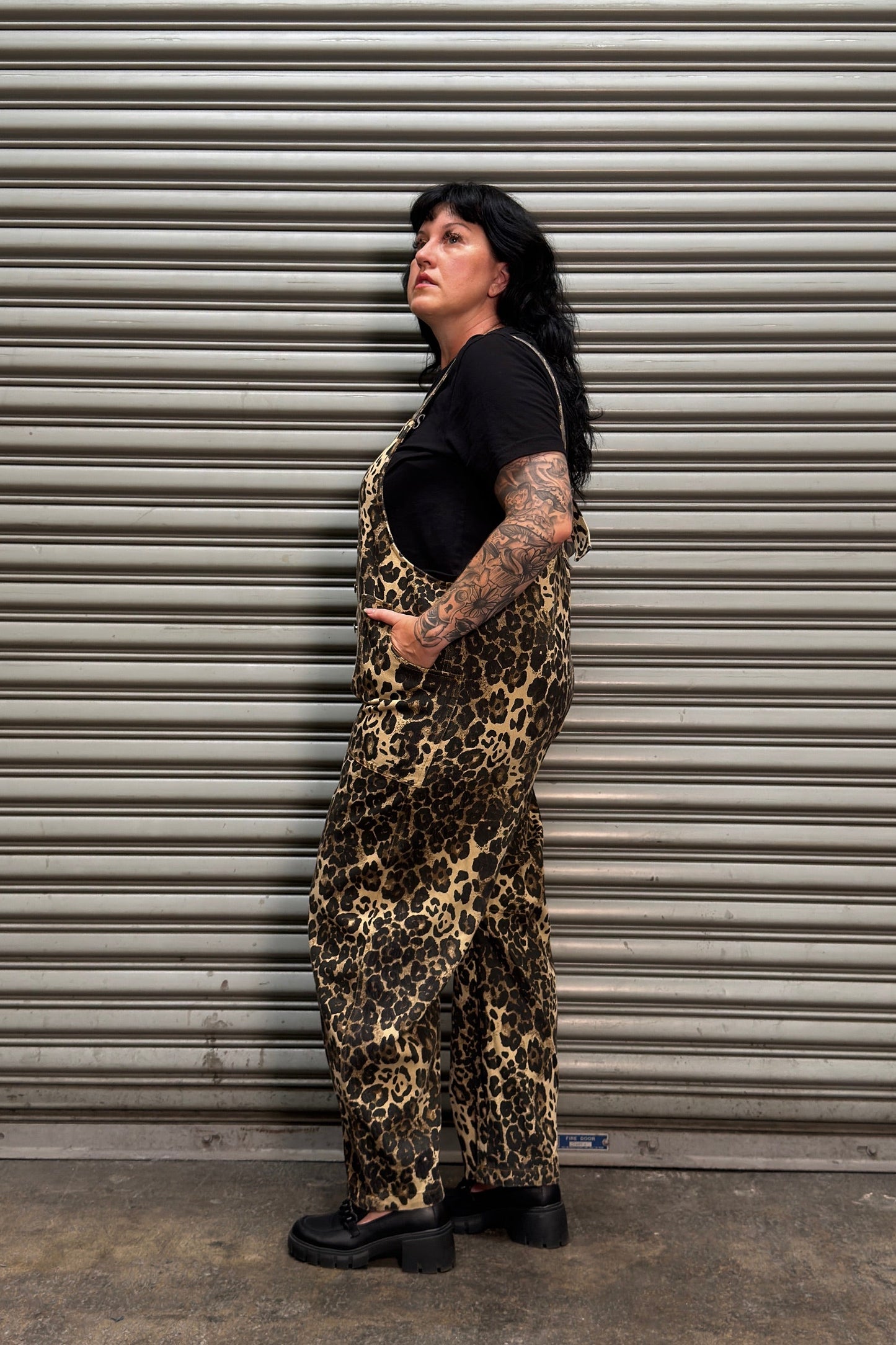 A person with dark hair and visible arm tattoos stands in profile in front of a gray metal roll-up door, wearing leopard-print overall-style shorts over a black T-shirt and black shoes.