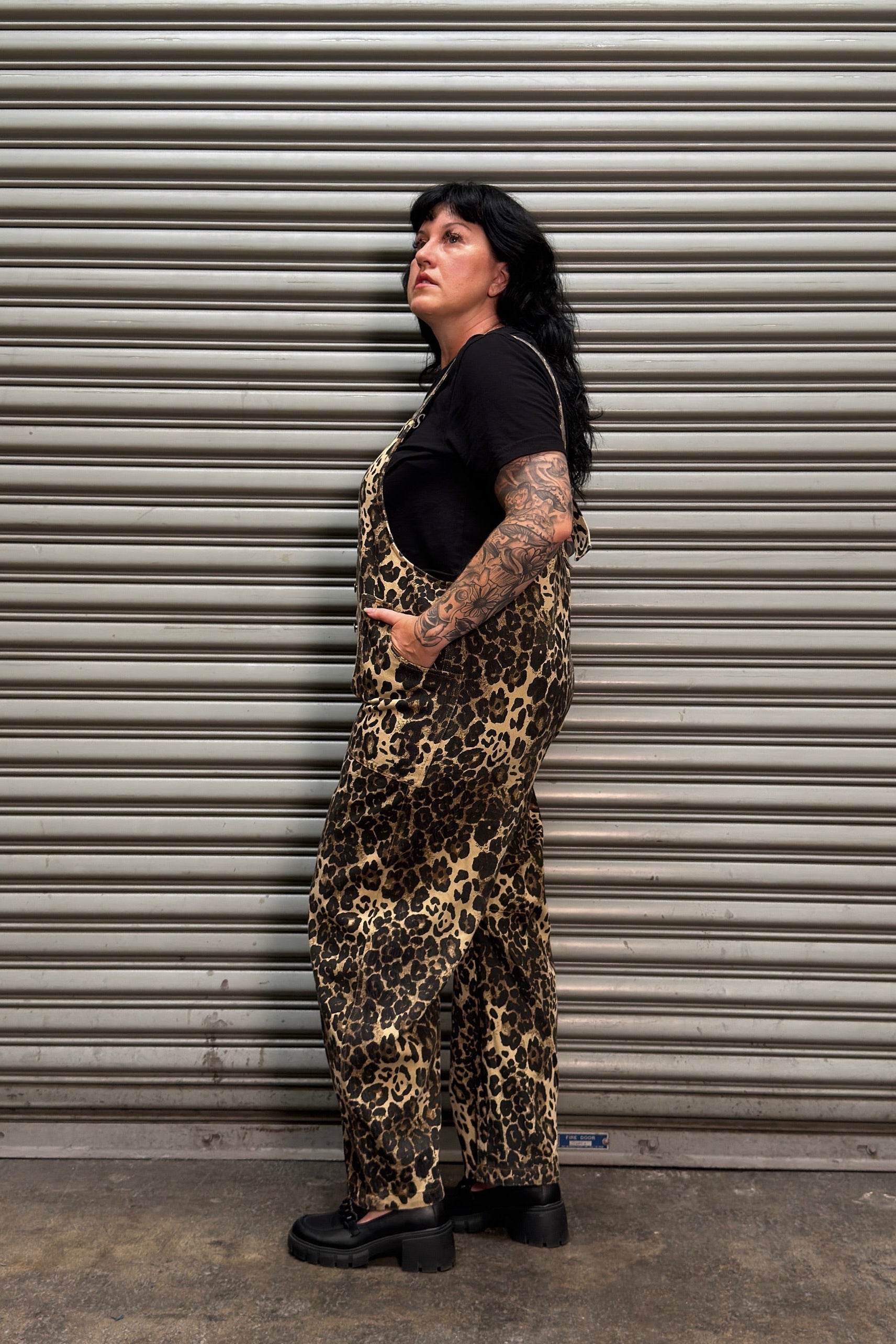 A person with dark hair and visible arm tattoos stands in profile in front of a gray metal roll-up door, wearing leopard-print overall-style shorts over a black T-shirt and black shoes.