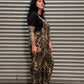 A woman with black hair and tattoos stands in front of a gray metal roll-up door, wearing a black short-sleeve top and brown-and-black leopard-print overall pants.