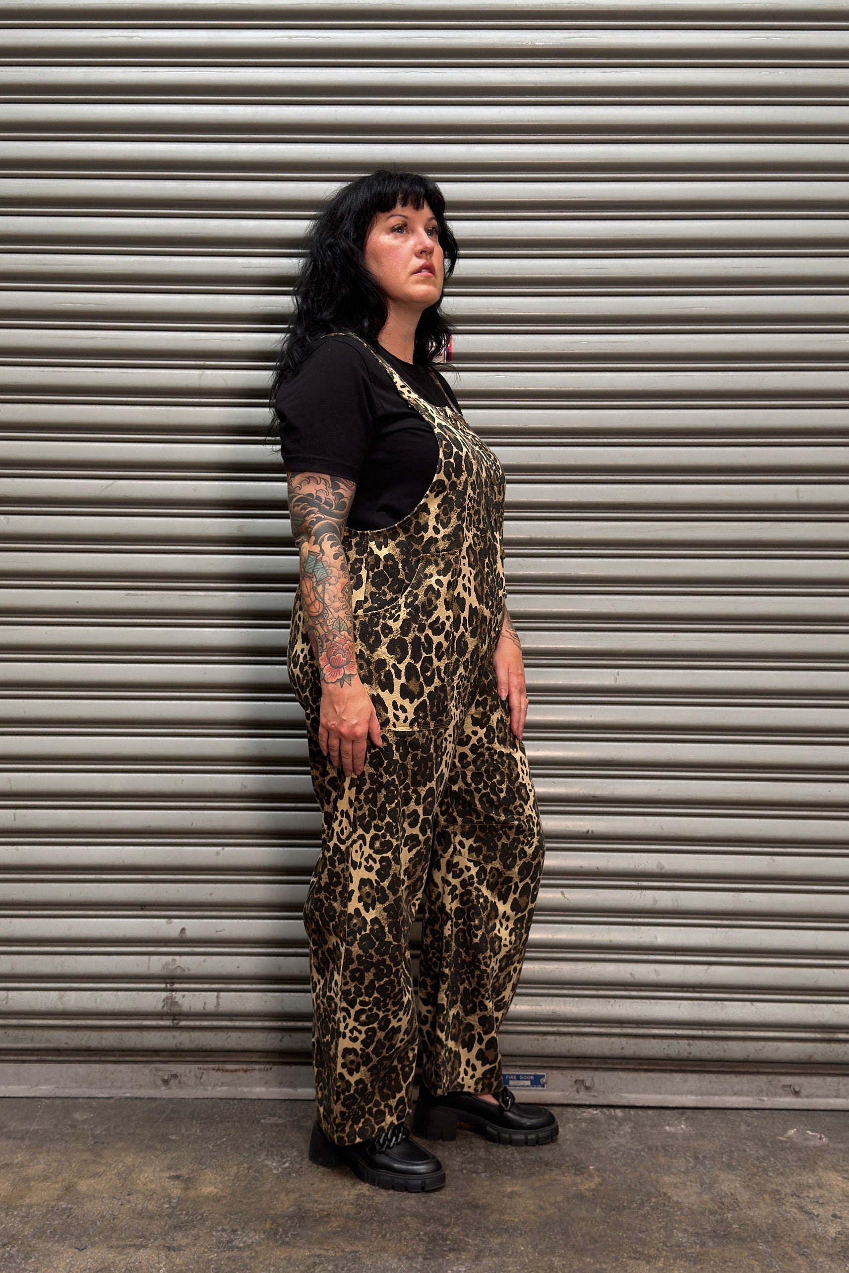 A woman with black hair and tattoos stands in front of a gray metal roll-up door, wearing a black short-sleeve top and brown-and-black leopard-print overall pants.