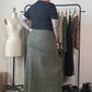 A person stands with their back to the camera in a room, wearing a black short-sleeve t-shirt and a long olive green and black leopard print midi skirt. A clothing rack with other garments, a dress form, and shoes are visible in the background.