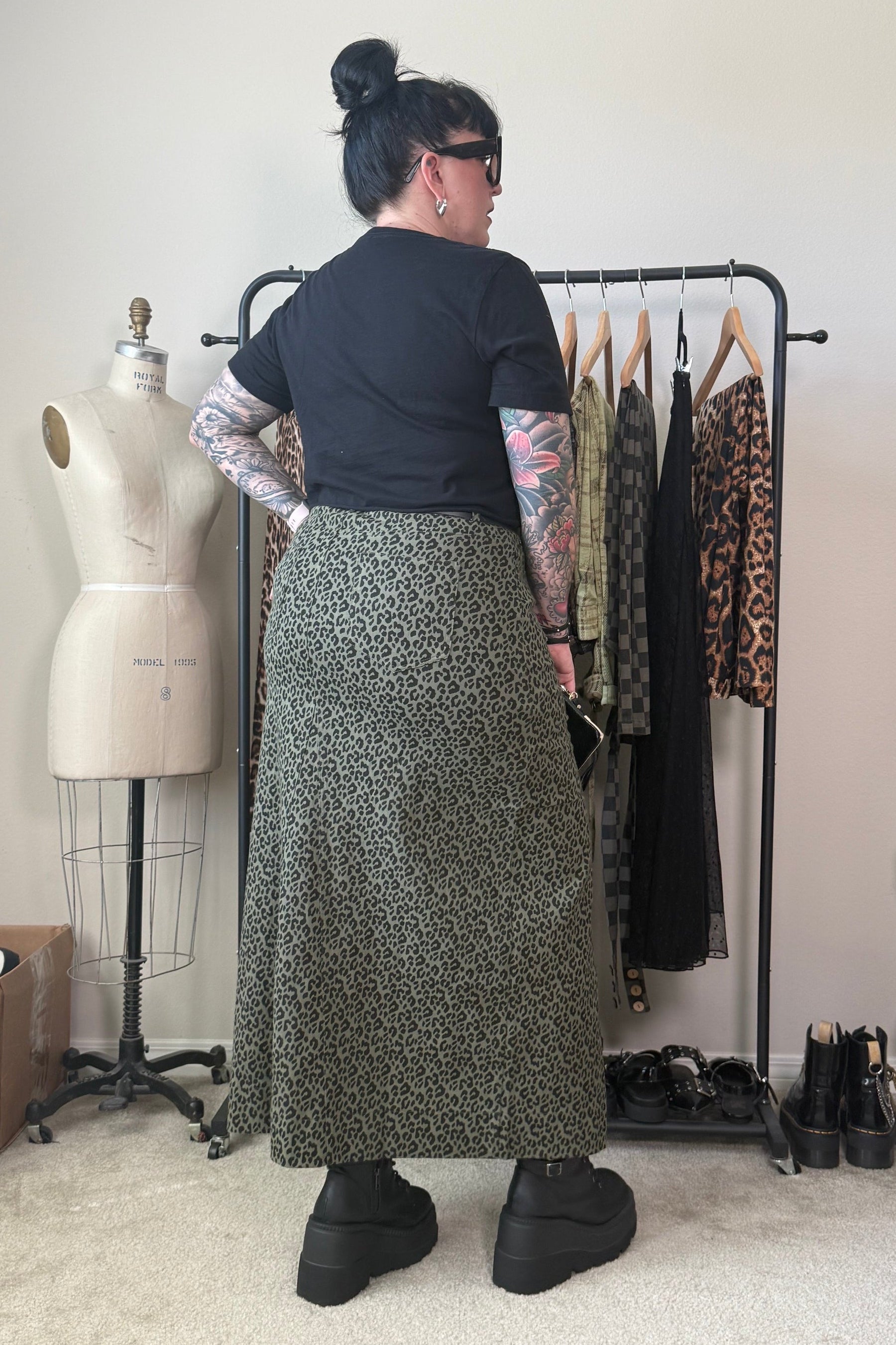 A person stands with their back to the camera in a room, wearing a black short-sleeve t-shirt and a long olive green and black leopard print midi skirt. A clothing rack with other garments, a dress form, and shoes are visible in the background.