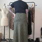 A person standing with their back to the camera in a room, wearing a black short-sleeve top and a long olive green and black leopard-print skirt. A clothing rack with leopard-print and black dresses, a dress form, and shoes are visible in the background.