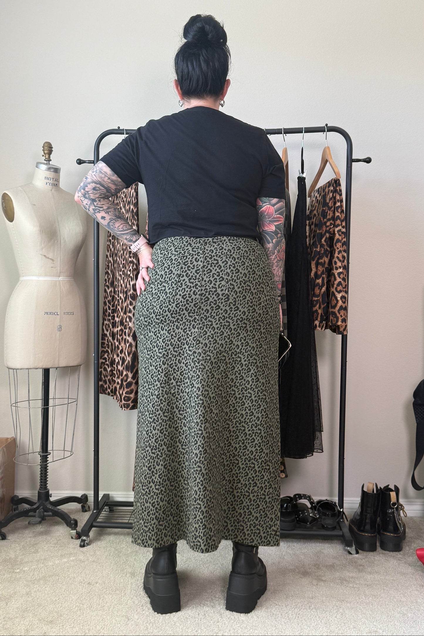 A person standing with their back to the camera in a room, wearing a black short-sleeve top and a long olive green and black leopard-print skirt. A clothing rack with leopard-print and black dresses, a dress form, and shoes are visible in the background.