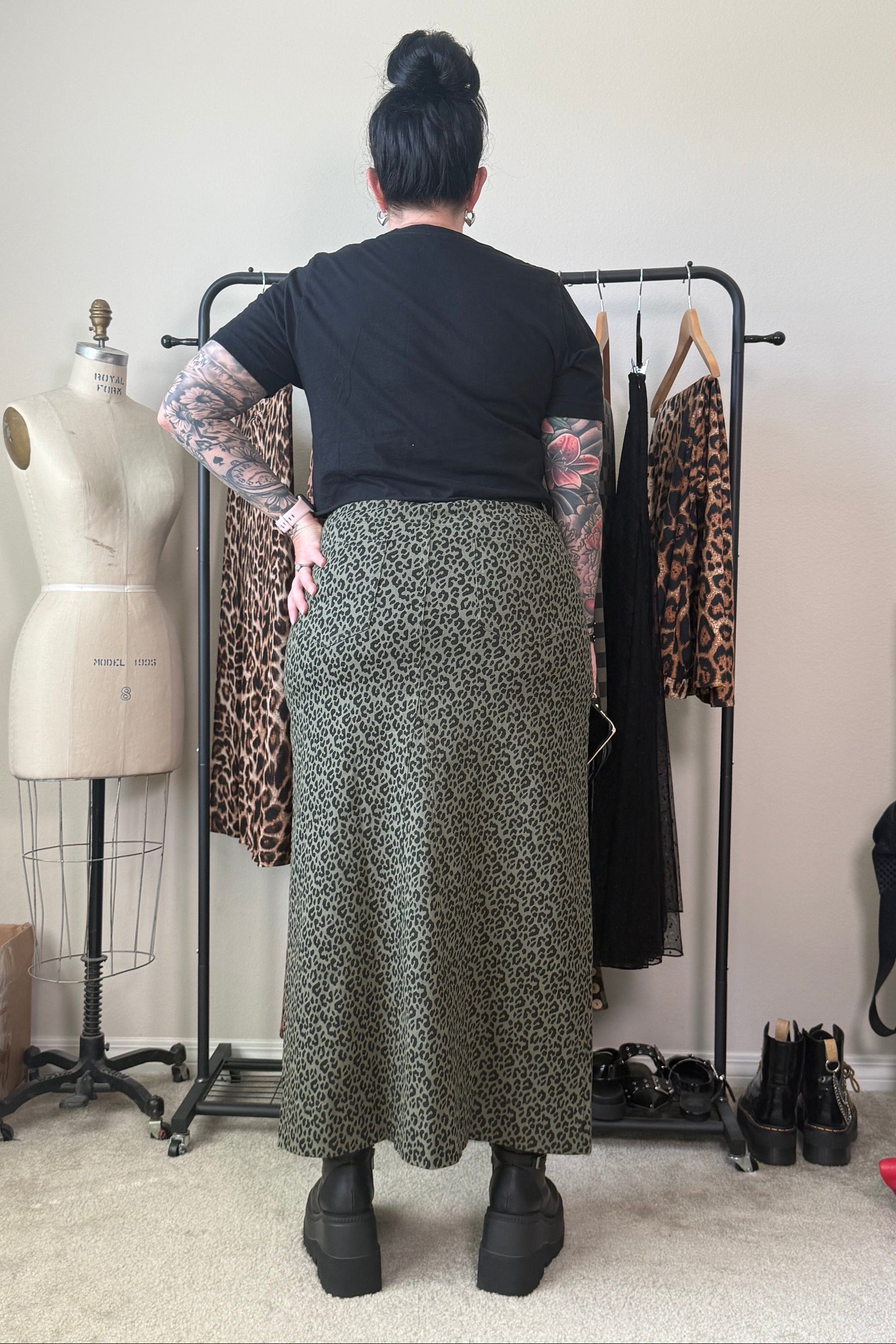 A person standing with their back to the camera in a room, wearing a black short-sleeve top and a long olive green and black leopard-print skirt. A clothing rack with leopard-print and black dresses, a dress form, and shoes are visible in the background.