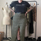 A person stands in a room wearing a black graphic T-shirt and a leopard-print skirt with a front slit, paired with black boots. A clothing rack, dress form, and shoes are visible in the background.
