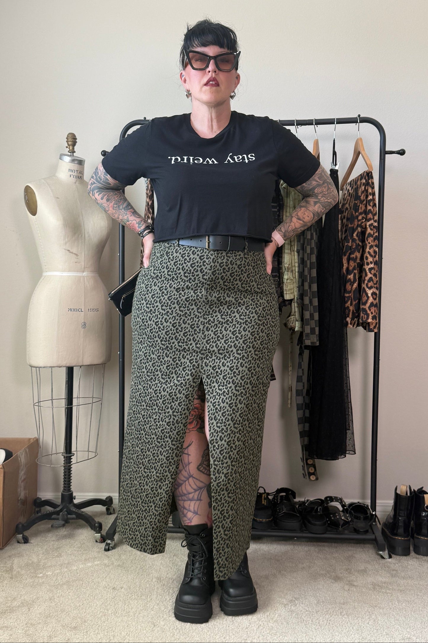 A person stands in a room wearing a black graphic T-shirt and a leopard-print skirt with a front slit, paired with black boots. A clothing rack, dress form, and shoes are visible in the background.