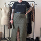 A person stands in a room wearing a black graphic T-shirt and a green leopard-print skirt with a front slit, paired with black boots. A clothing rack with patterned garments, a dress form, and shoes are visible in the background.