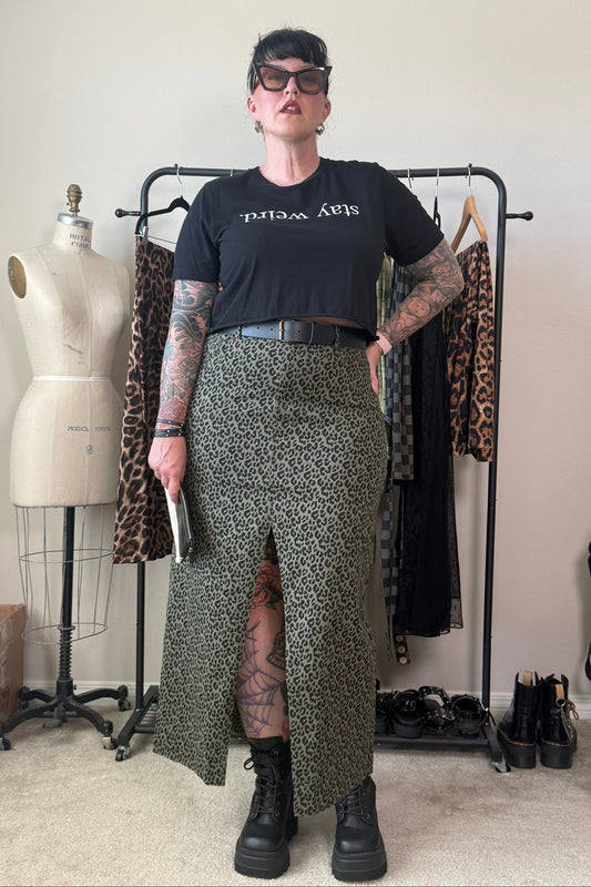 A person stands in a room wearing a black graphic T-shirt and a green leopard-print skirt with a front slit, paired with black boots. A clothing rack with patterned garments, a dress form, and shoes are visible in the background.