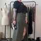 A person stands in a room wearing a black short-sleeve top and a long olive green leopard-print skirt with a thigh slit. They are wearing black platform boots and sunglasses, standing in front of a clothing rack with other garments and a dress form mannequin.