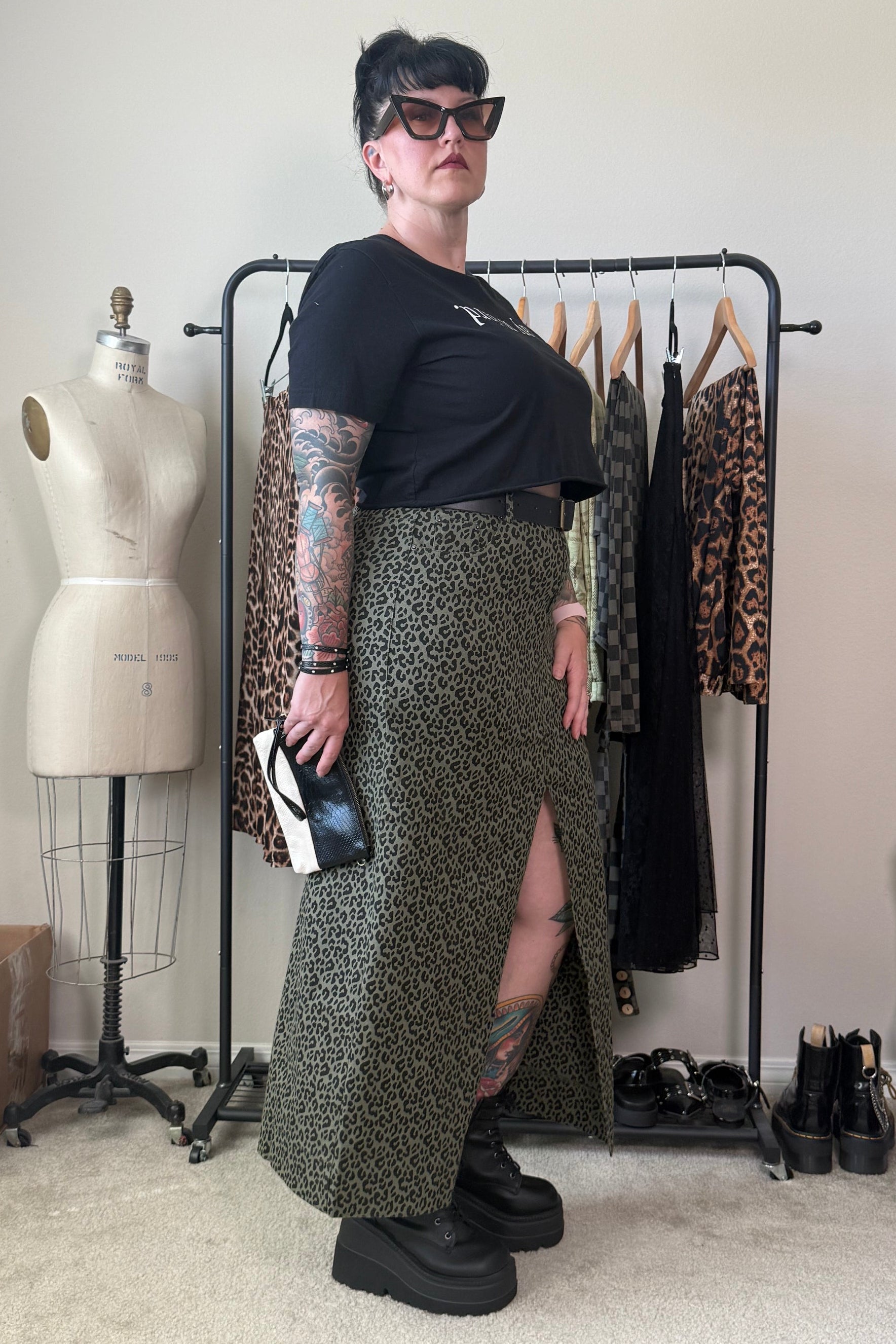A person stands in a room wearing a black short-sleeve top and a long olive green leopard-print skirt with a thigh slit. They are wearing black platform boots and sunglasses, standing in front of a clothing rack with other garments and a dress form mannequin.