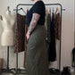 A person stands in profile wearing a black short-sleeve top and a long olive green leopard-print skirt, with black platform boots, in a room with a clothing rack, mannequin, and shoes.