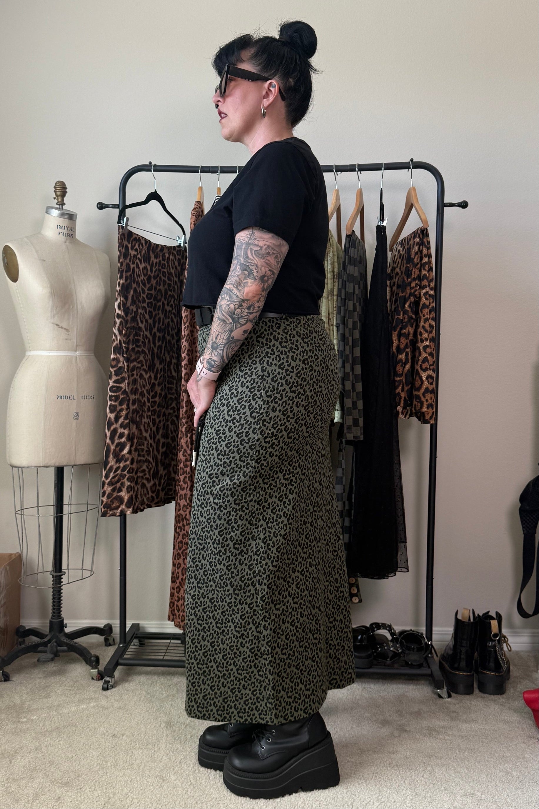 A person stands in profile wearing a black short-sleeve top and a long olive green leopard-print skirt, with black platform boots, in a room with a clothing rack, mannequin, and shoes.