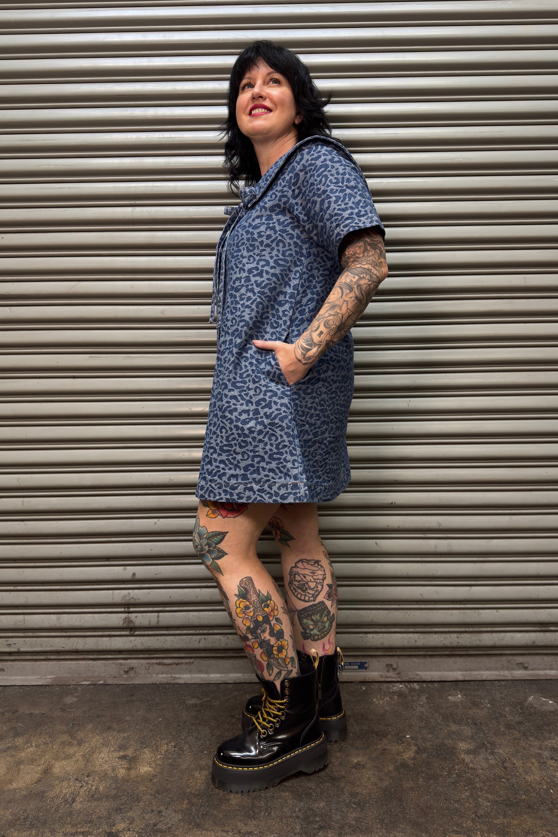 Person wearing a blue patterned dress and black boots standing against a corrugated metal wall.